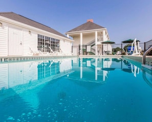 A swimming pool with a clear blue water, a white building with a covered patio, and a staircase leading to a second floor.