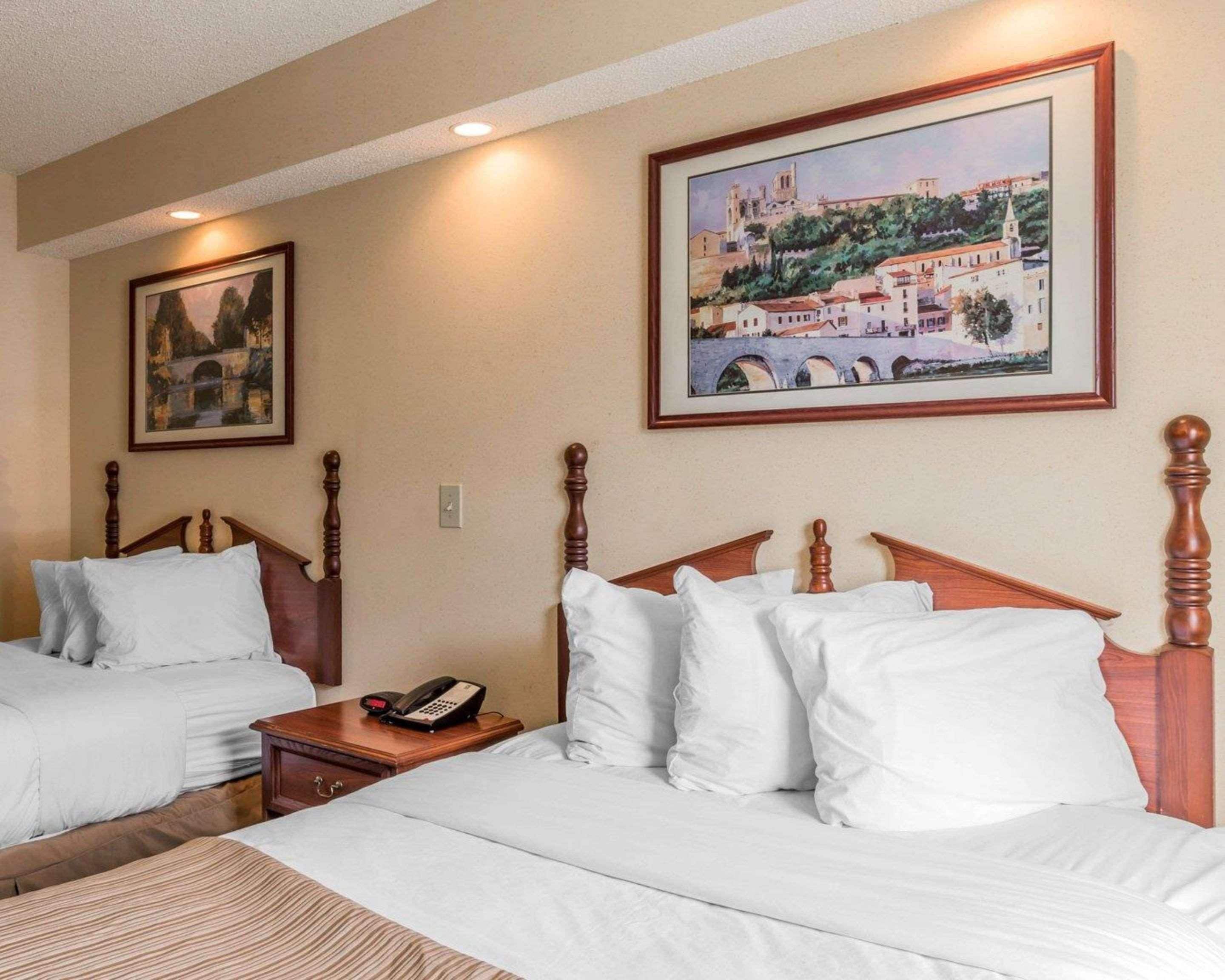 A hotel room with two beds, a wooden headboard, a bedside table with a phone, and two framed pictures on the wall.