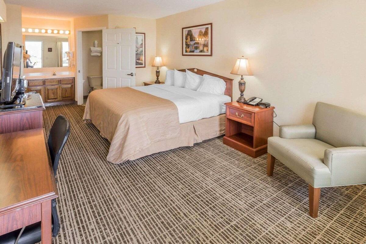 Habitación de hotel con cama, mesita de noche, lámpara, silla, escritorio, televisor y baño con espejo e iluminación.