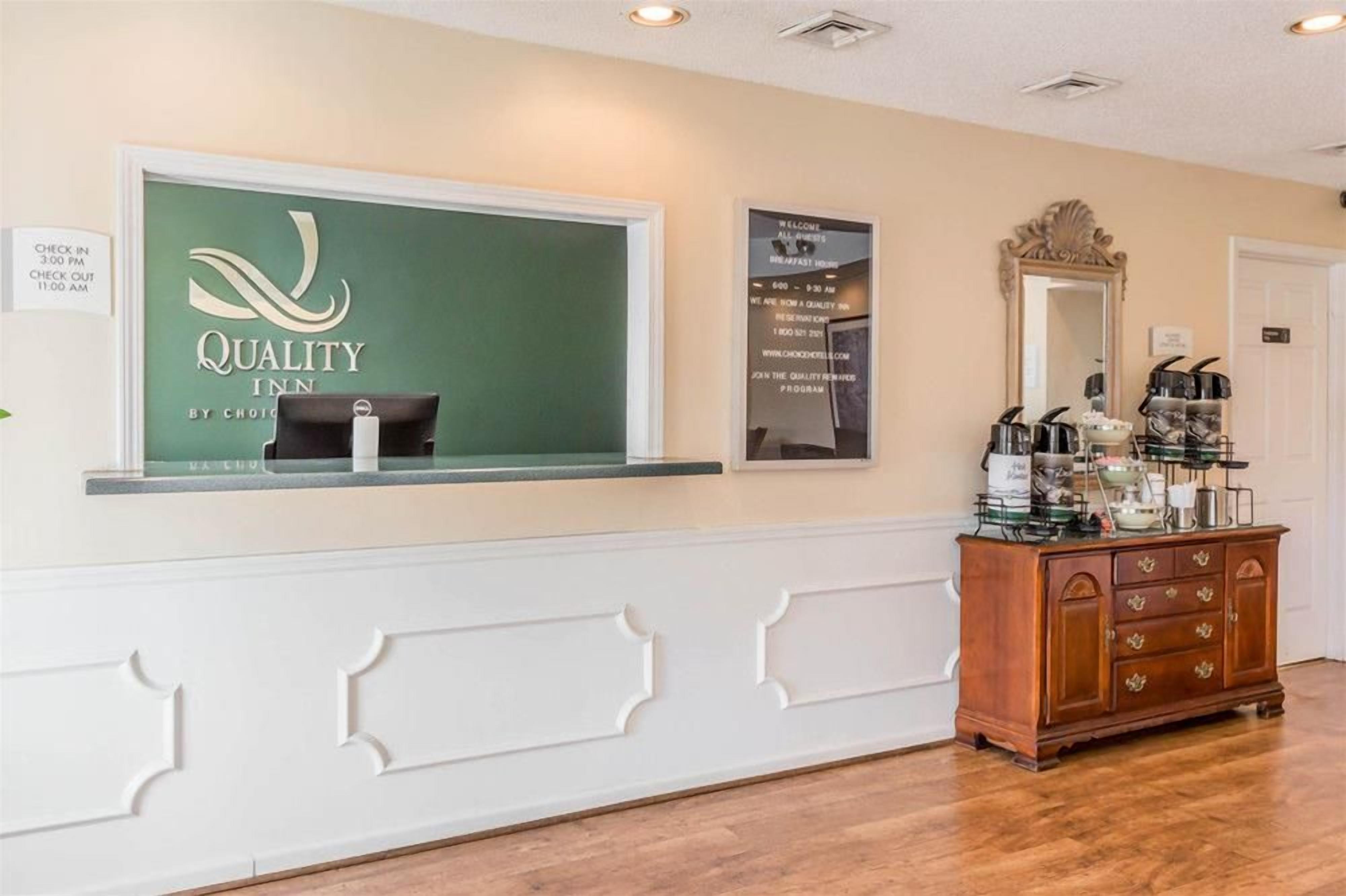 Un espace d’accueil avec un panneau indiquant « Quality Inn », un tableau affichant les heures d’arrivée et de départ, un miroir et un coin café.