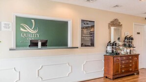 A reception area with a 'Quality Inn' sign, a check-in/out schedule, a mirror, and a coffee station.