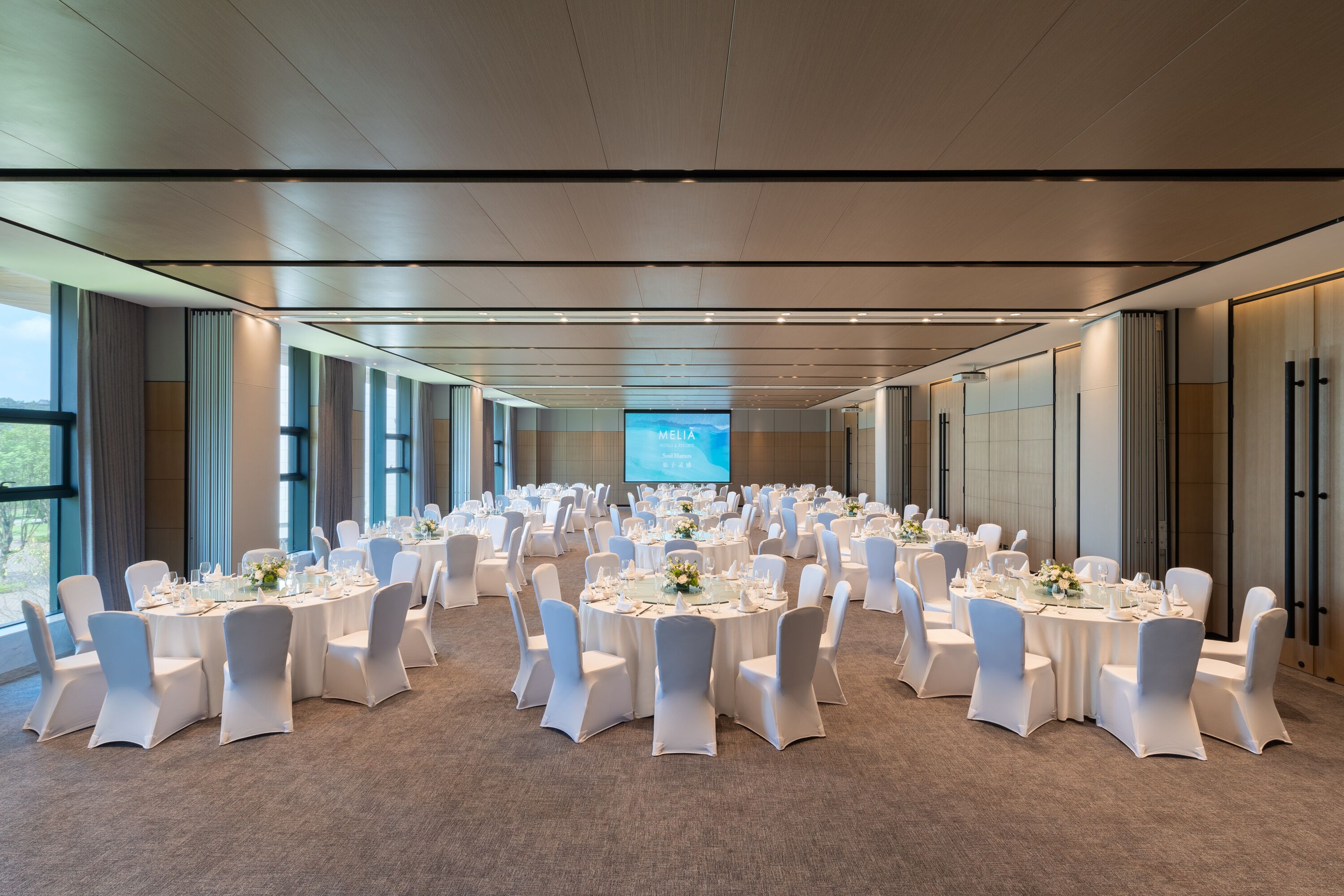 Une grande salle de conférence avec des tables rondes et des chaises blanches, un grand écran à l’avant et de grandes fenêtres le long d’un des murs.