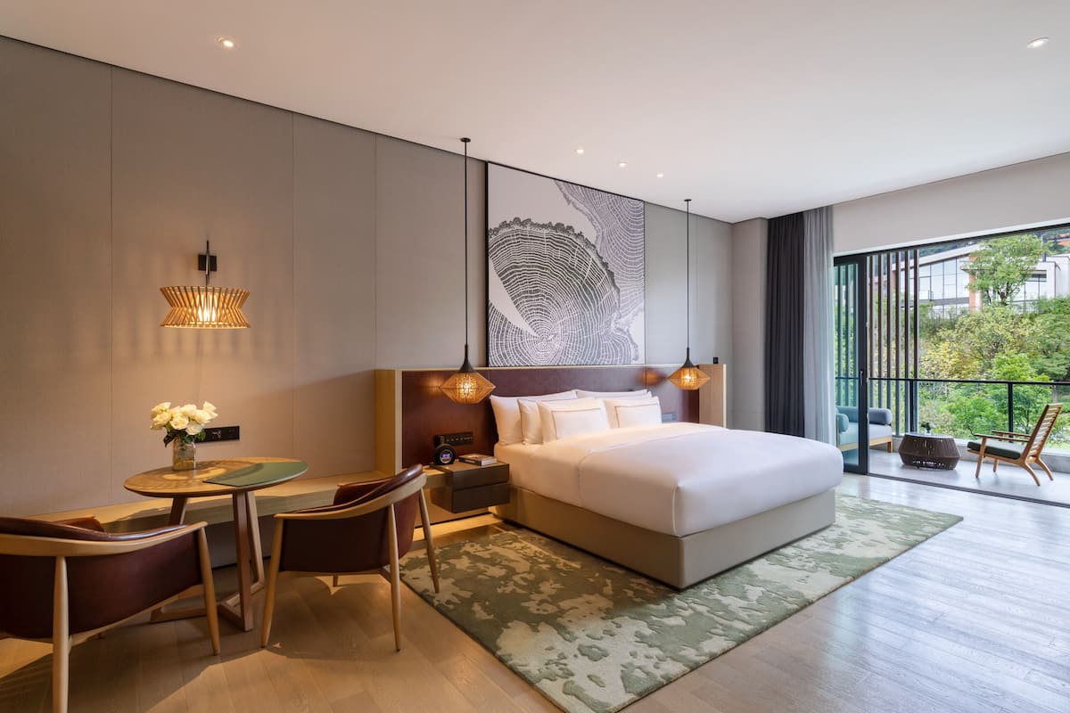 A modern hotel room with a large bed, a desk with a chair, a small table with flowers, and a large window with a view of greenery.