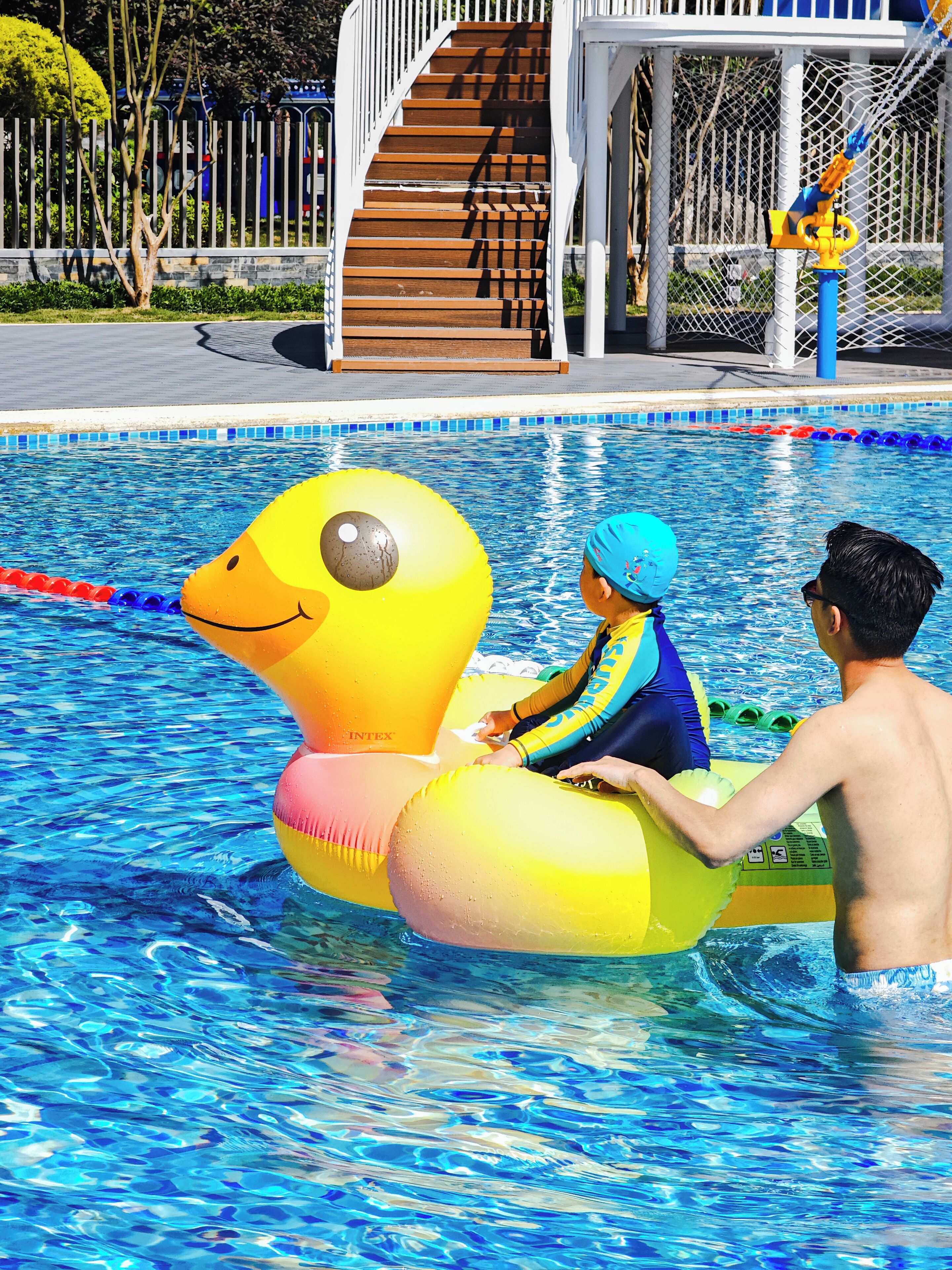 Ein Schwimmbad mit einer Enten-Schwimmweste, einem Kind darauf und einem Mann neben dem Pool.