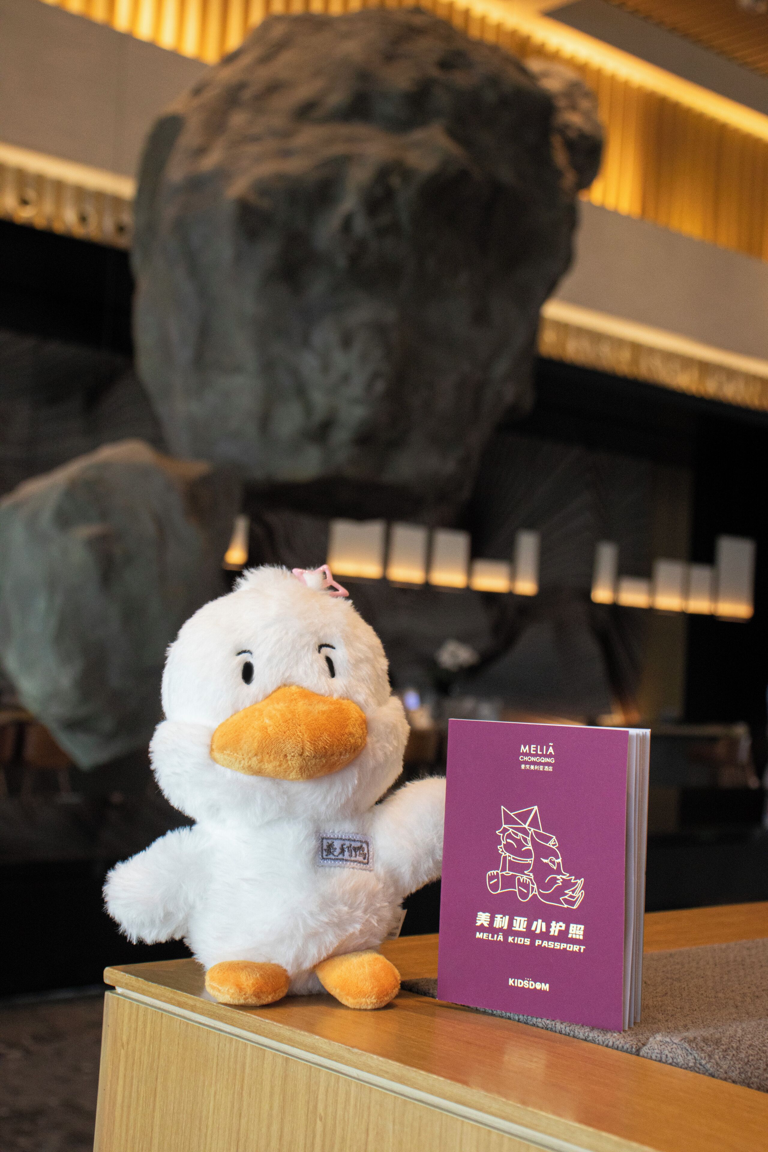 Un patito de peluche sobre una superficie de madera, con un folleto morado etiquetado como 'MELIA KIDS PASSPORT' a su lado.