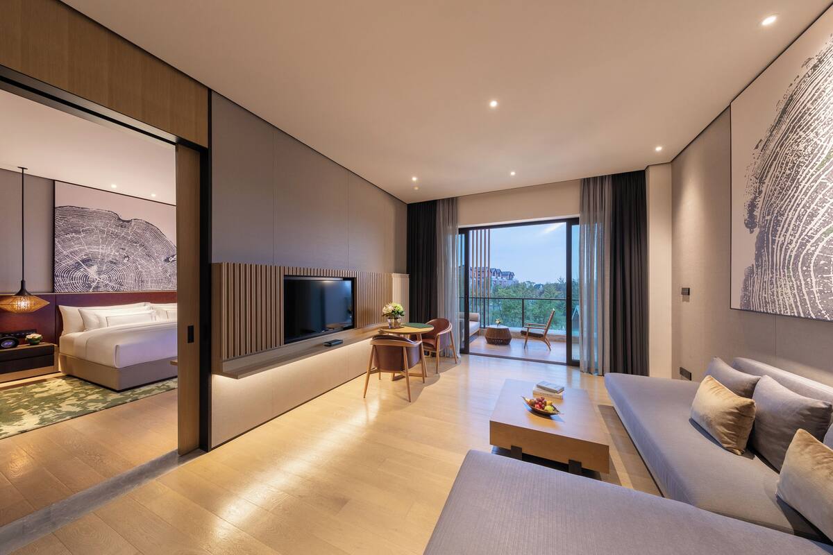 A modern hotel room with a bed, TV, sofa, dining table, and a balcony with a view.