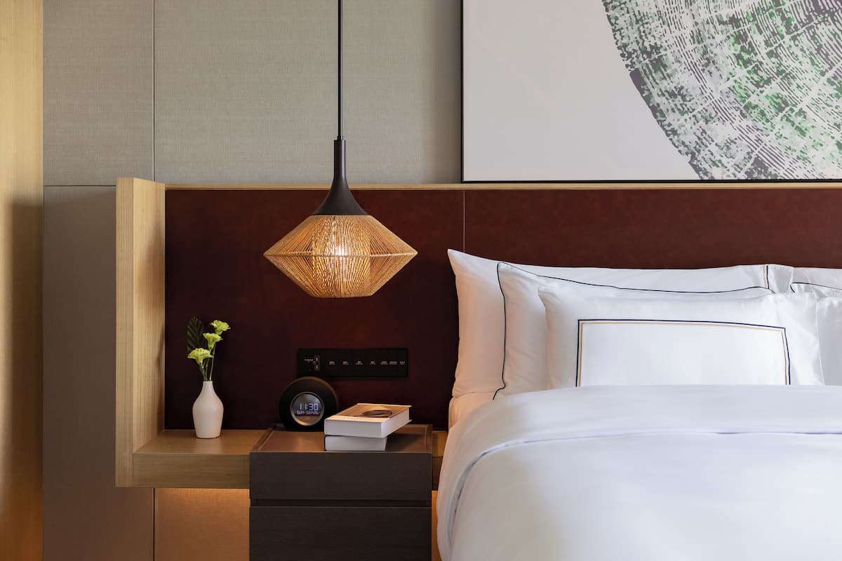 A modern hotel room with a bed, bedside table, lamp, clock, and a vase with flowers.