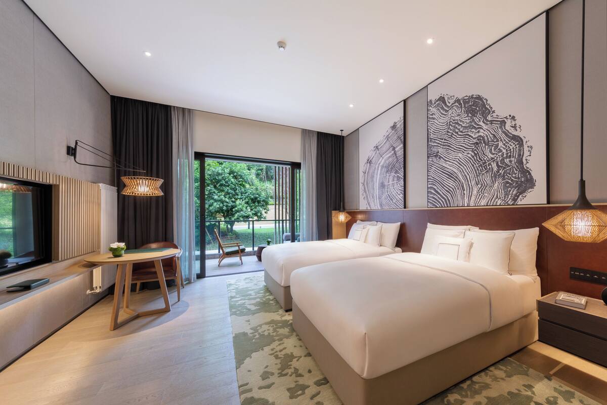 A modern hotel room with two beds, a desk, a chair, and a large window offering a view of greenery.