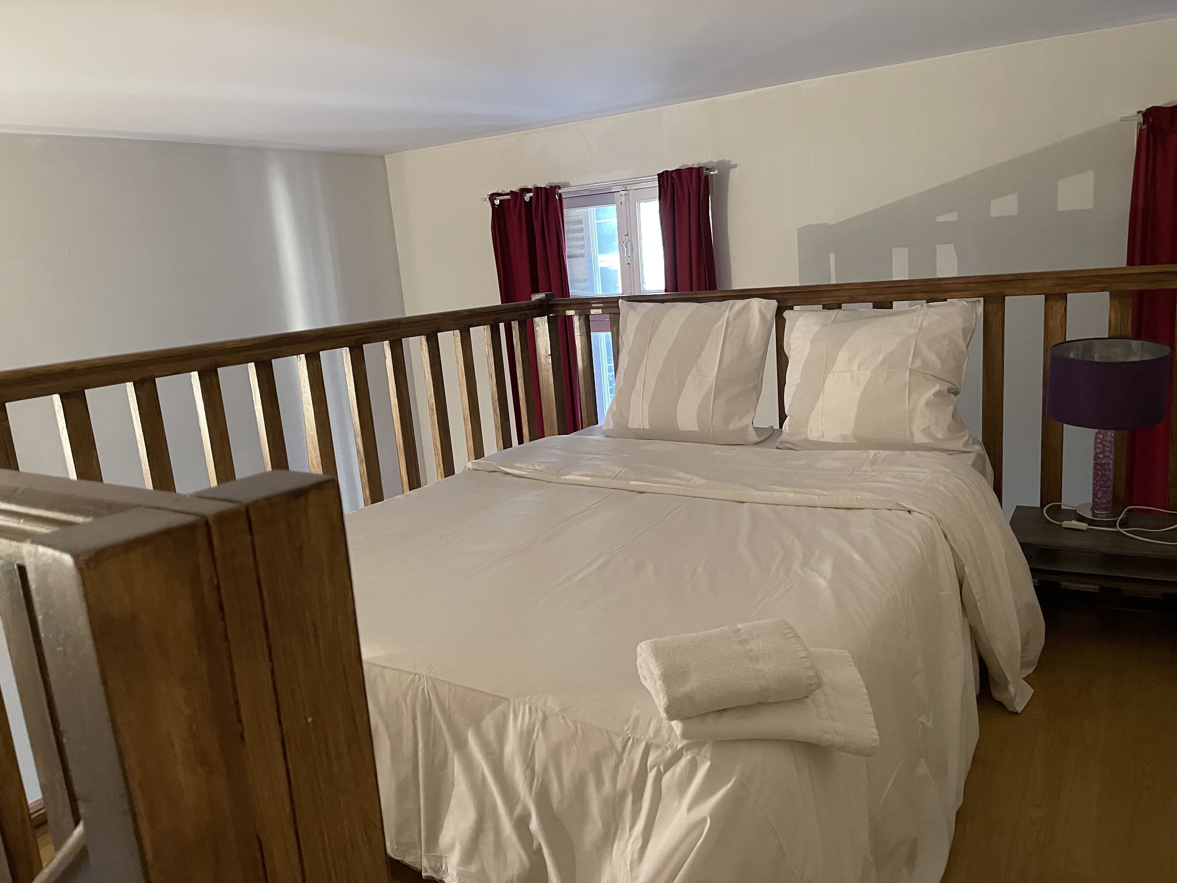 A neatly made bed with white linens and a wooden headboard in a room with a wooden floor and a window with red curtains.