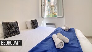Un dormitorio con una cama grande, una colcha azul, dos toallas enrolladas y una ventana con vistas abiertas a edificios.