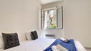 A bedroom with a large bed, a blue bedspread, two rolled towels, and a window with an open view of buildings.