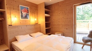 A wooden cabin bedroom with a bed, shelves, a small table, and a door leading to an outdoor area.
