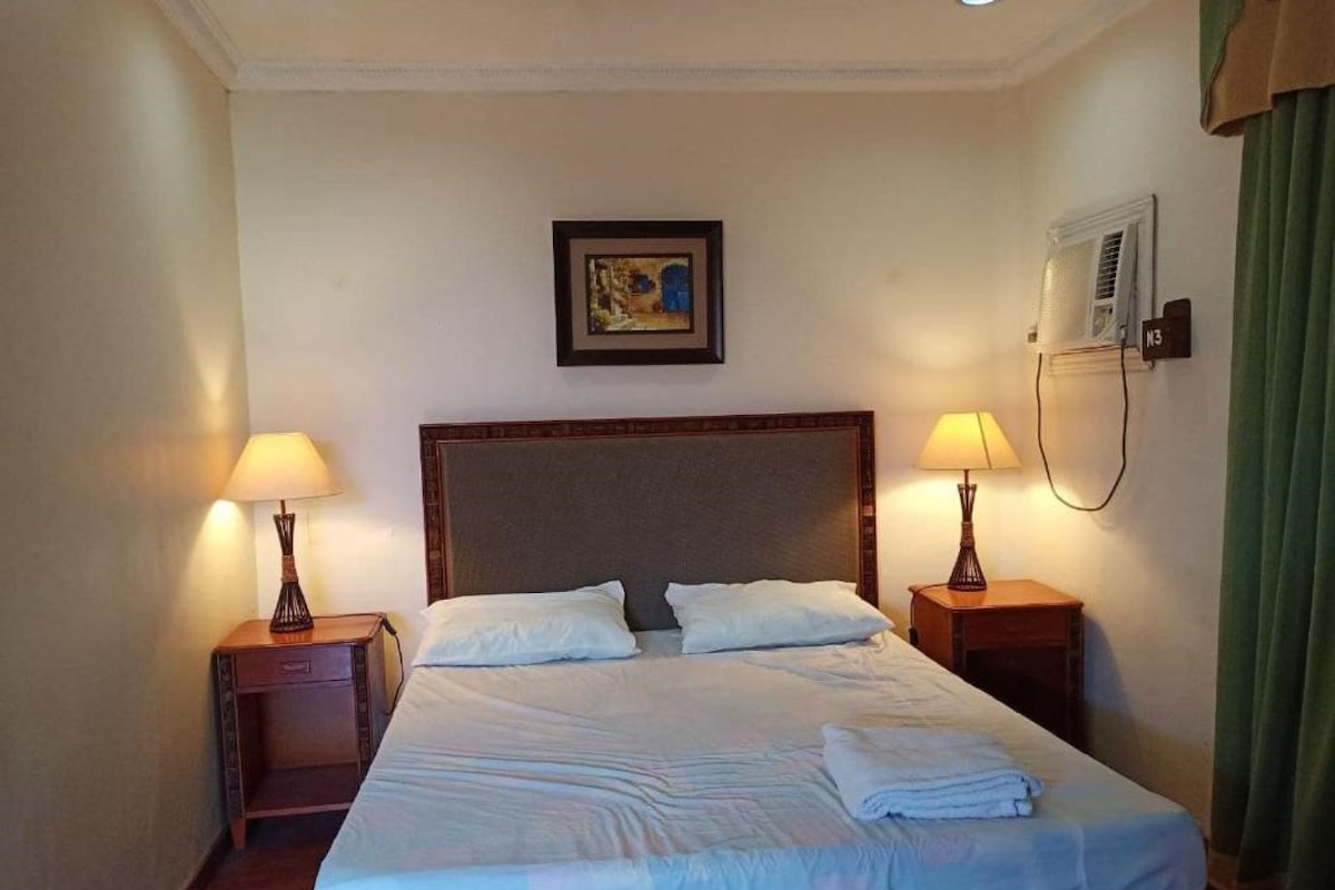 A hotel room with a large bed, two bedside tables with lamps, a framed picture on the wall, and a window with curtains.