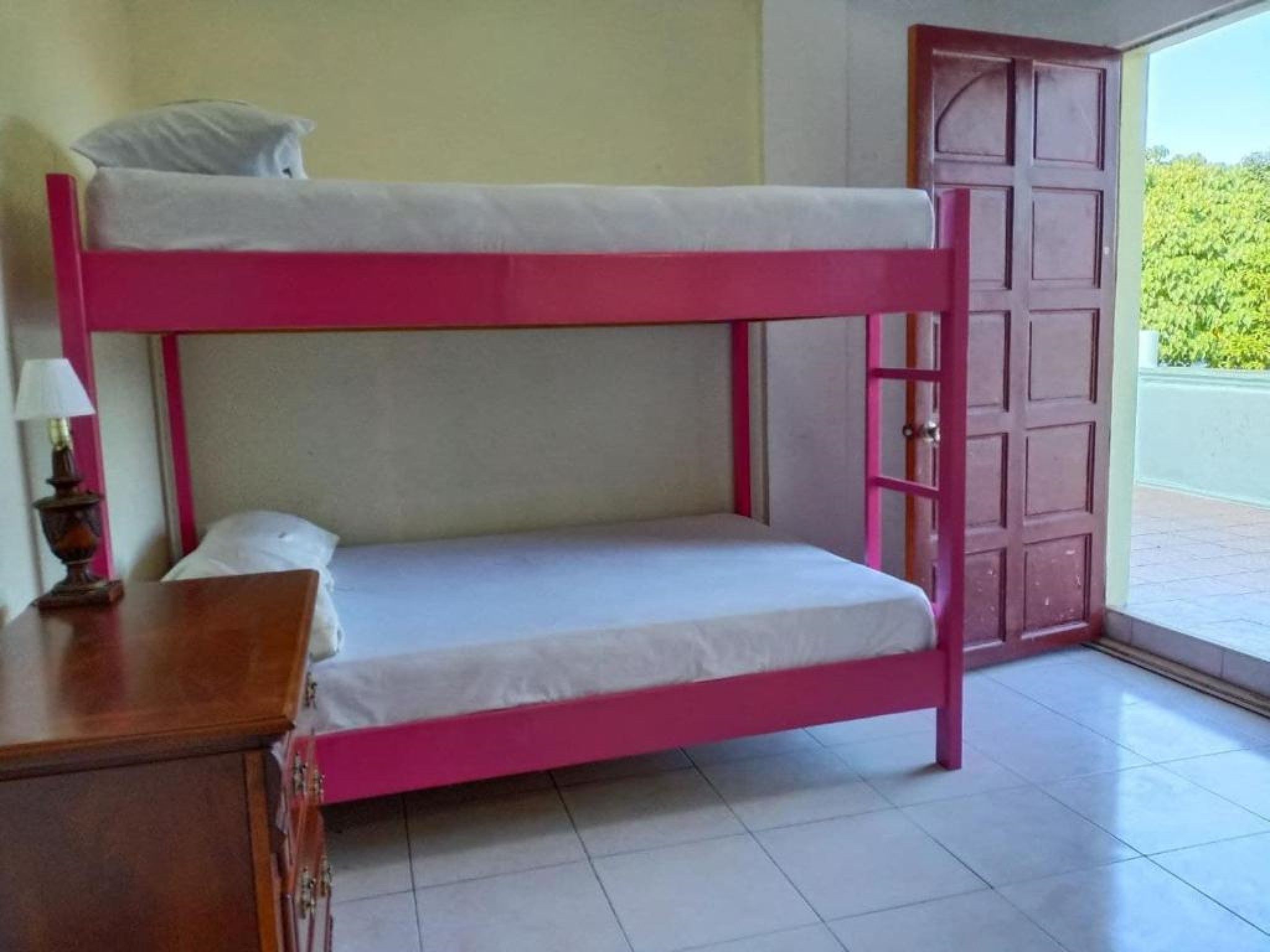 Una cama litera con estructura rosa y ropa de cama blanca, una mesita de noche de madera con una lámpara, y una puerta que da al exterior.