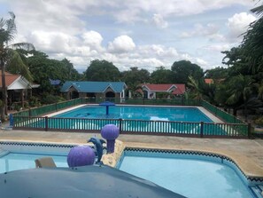 Une piscine entourée d’une clôture verte, bordée de palmiers et de bâtiments aux toits rouges.
