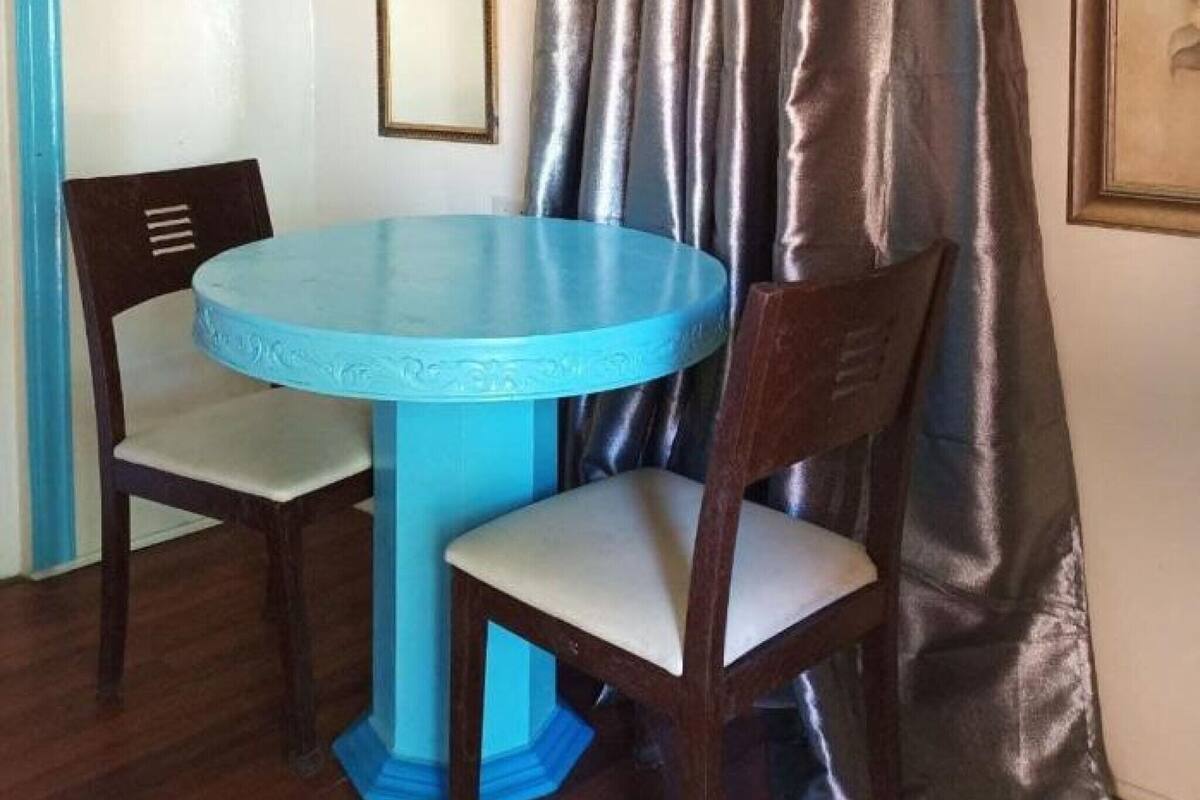 A room with a round blue table, two wooden chairs, a mirror on the wall, and a curtain.