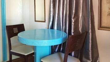 A room with a round table and two chairs, a blue base, a wooden chair with a cushion, a framed picture on the wall, and a curtain.