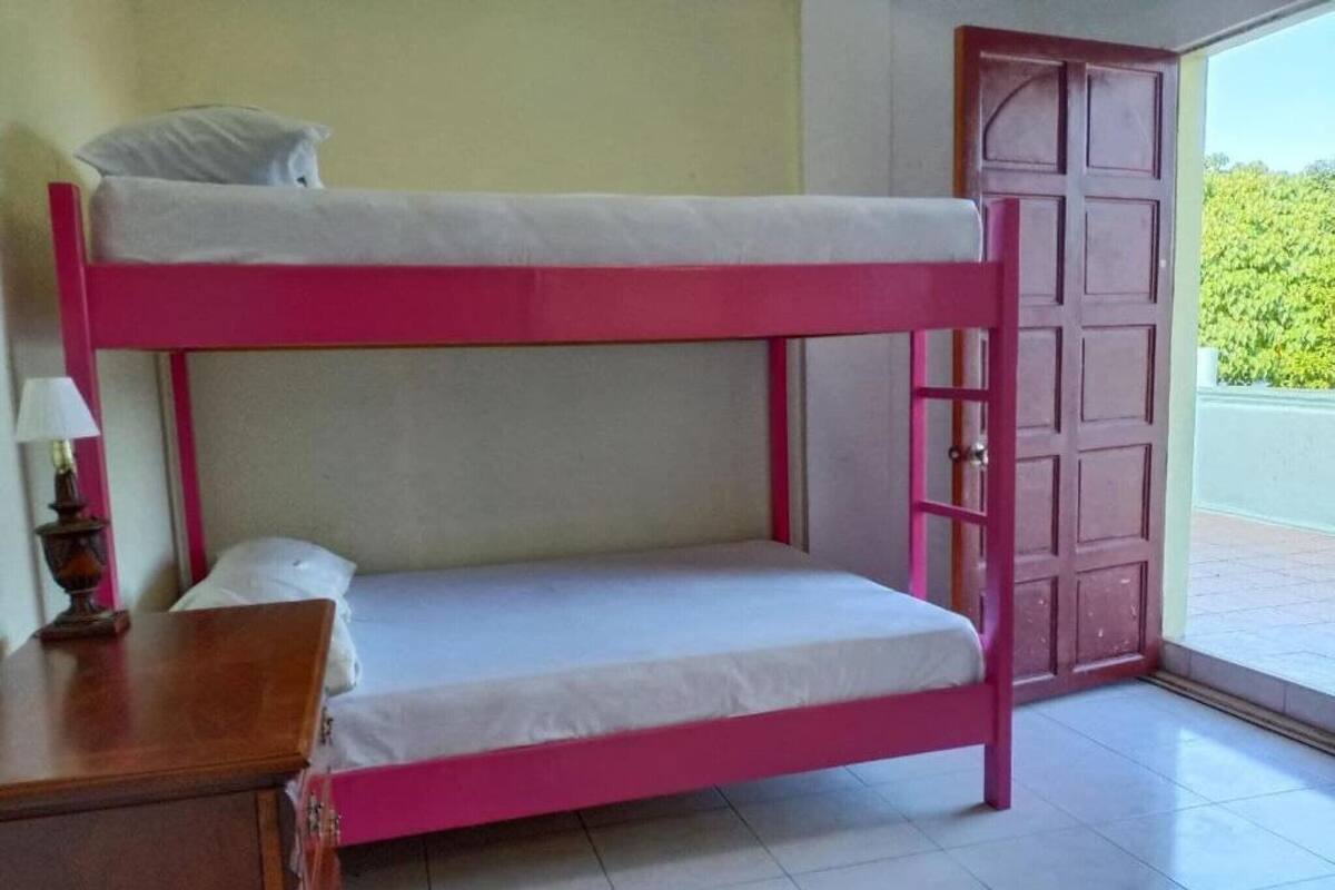 A bunk bed with a pink frame and white bedding, a wooden nightstand with a lamp, and a door leading outside.