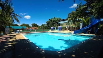 A swimming pool with a blue slide, surrounded by palm trees and a building with a green roof.