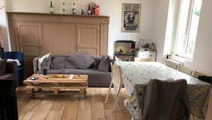 Ein gemütliches Wohnzimmer mit einer Couch, einem Holztisch und einem Esstisch mit Stühlen. Ein Fernseher und ein Poster sind an der Wand angebracht.
