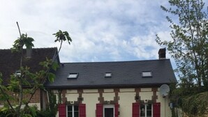 Ein zweistöckiges Haus mit geneigtem Dach, roten Fensterläden und einem kleinen Vorgarten mit einer Bank.