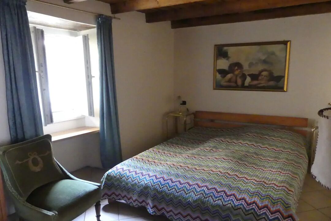 A bedroom with a bed, a chair, a window with curtains, a nightstand, and a painting on the wall.
