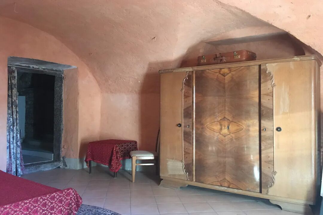 A room with a wooden wardrobe, a small table with a red cloth, a chair, and a bed with a patterned cover.