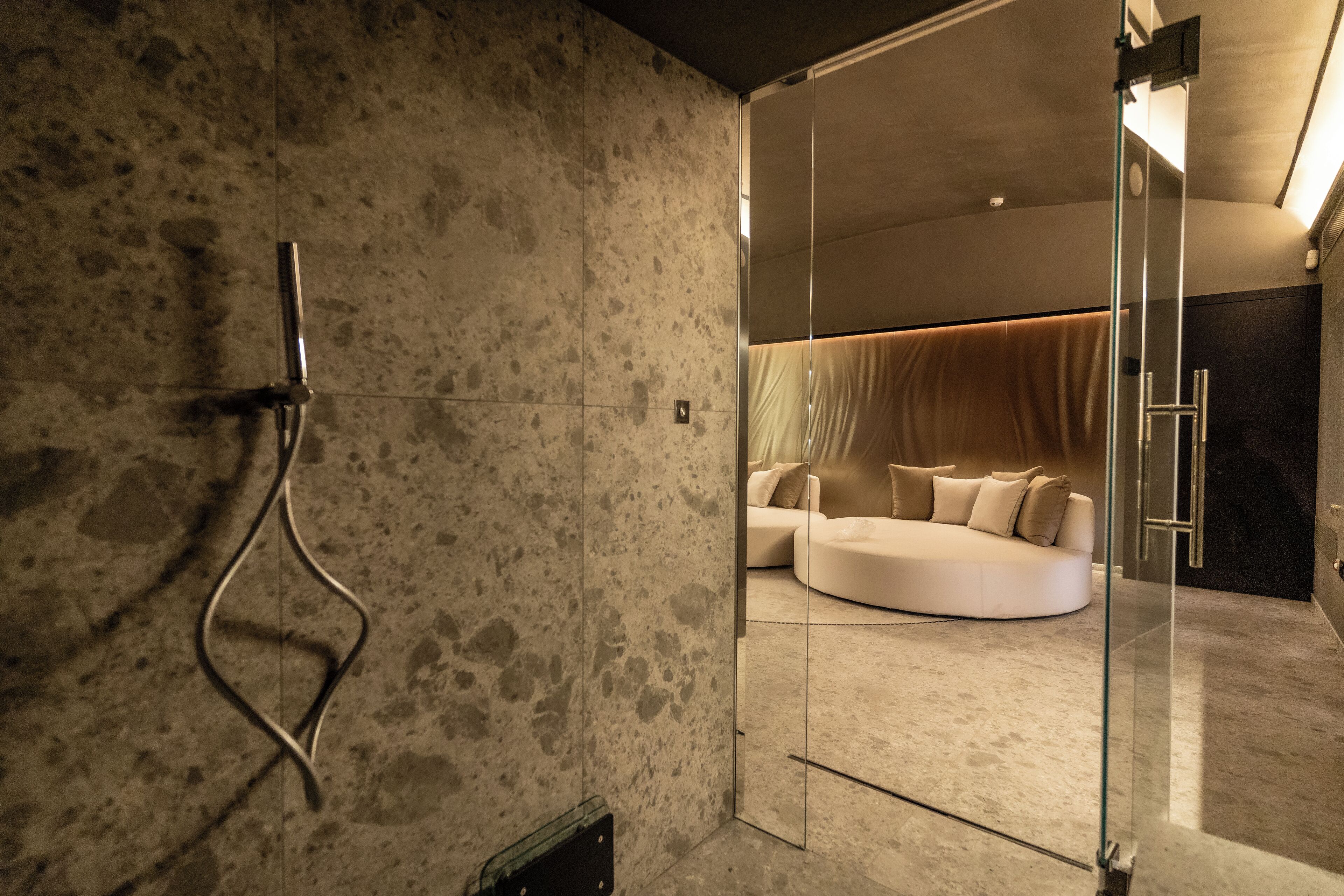 A modern bathroom with a glass shower enclosure, a curved white sofa in the background, and a textured wall.