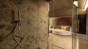 A modern bathroom with a glass shower enclosure, a curved white sofa in the background, and a textured wall.