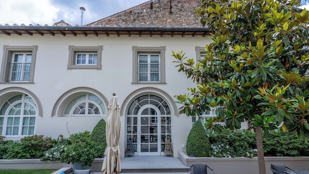 Un edificio con fachada blanca, varios ventanales de arco y un jardín cuidado con mesas al aire libre.
