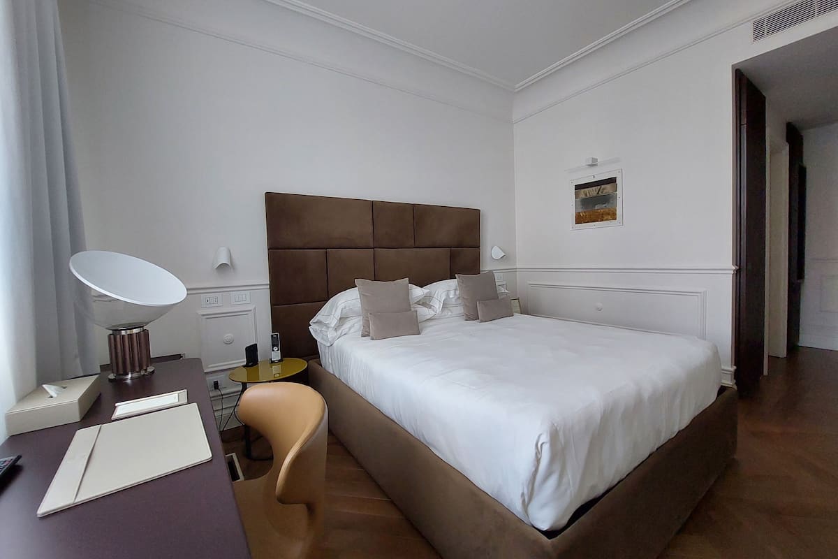 A hotel room with a large bed, a desk with a lamp, a chair, and a small table.