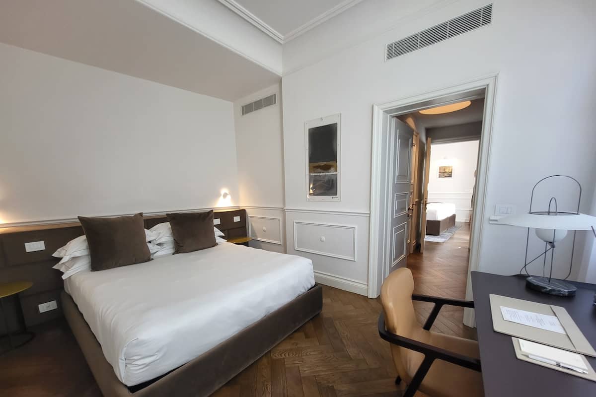 A hotel room with a bed, a desk, a chair, a lamp, and a door leading to another room.