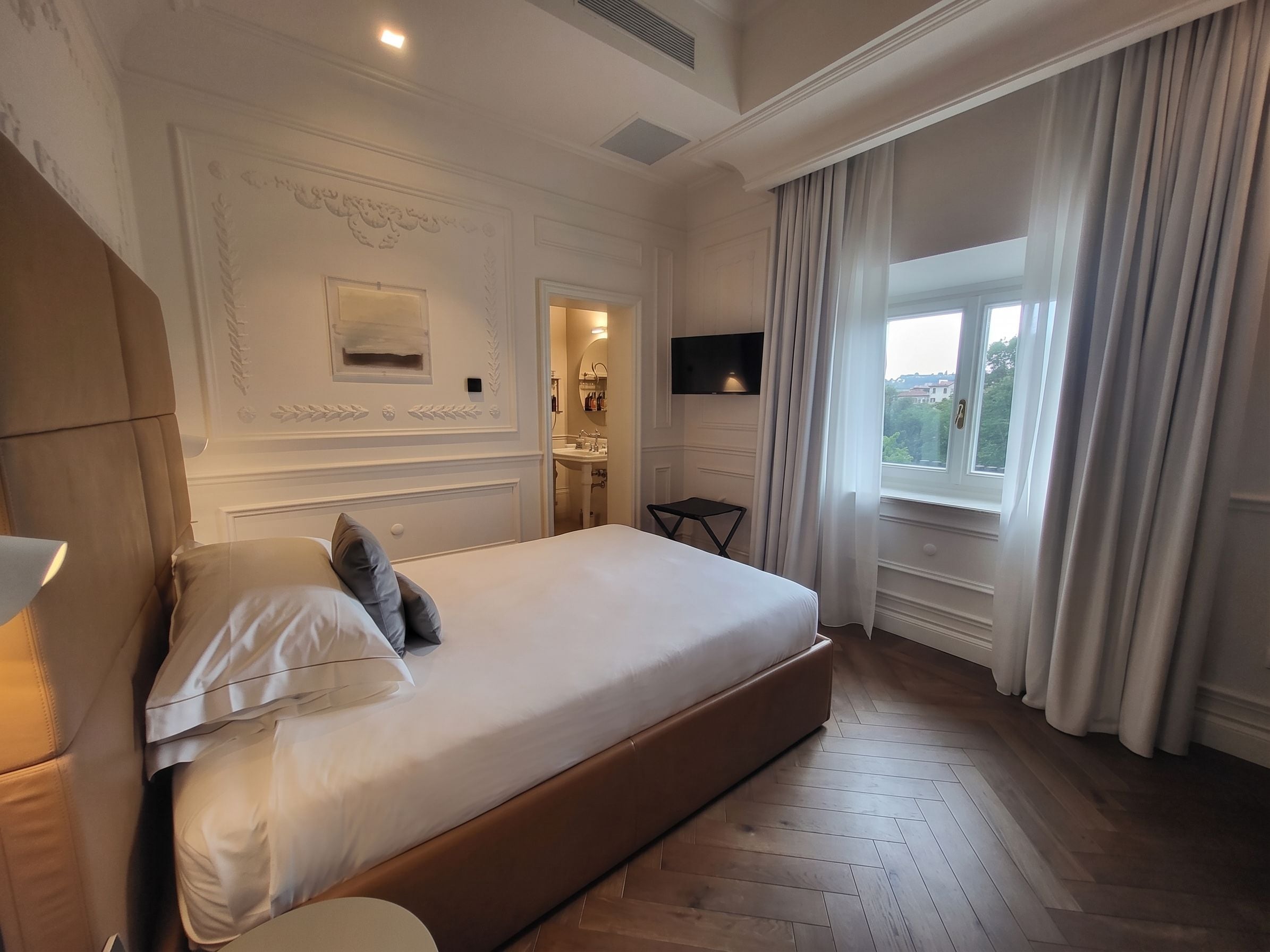 Habitación de hotel con una cama grande, una mesita de noche, un televisor y un ventanal con cortinas.