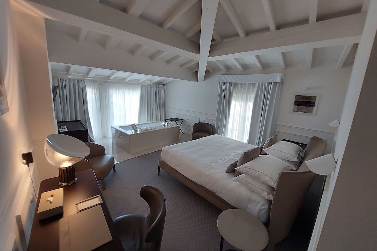A hotel room with a large bed, a desk, a chair, a small table, a desk lamp, and a window with curtains.