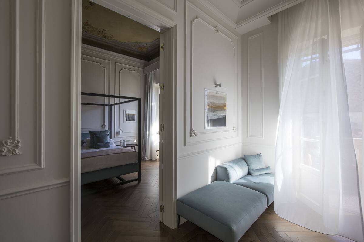A bedroom with a bed, a bench, a painting, and a window with sheer curtains.