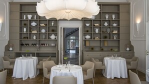 Comedor con dos mesas redondas, cada una rodeada por cuatro sillas, y una lámpara de araña grande y ornamentada sobre ellas.
