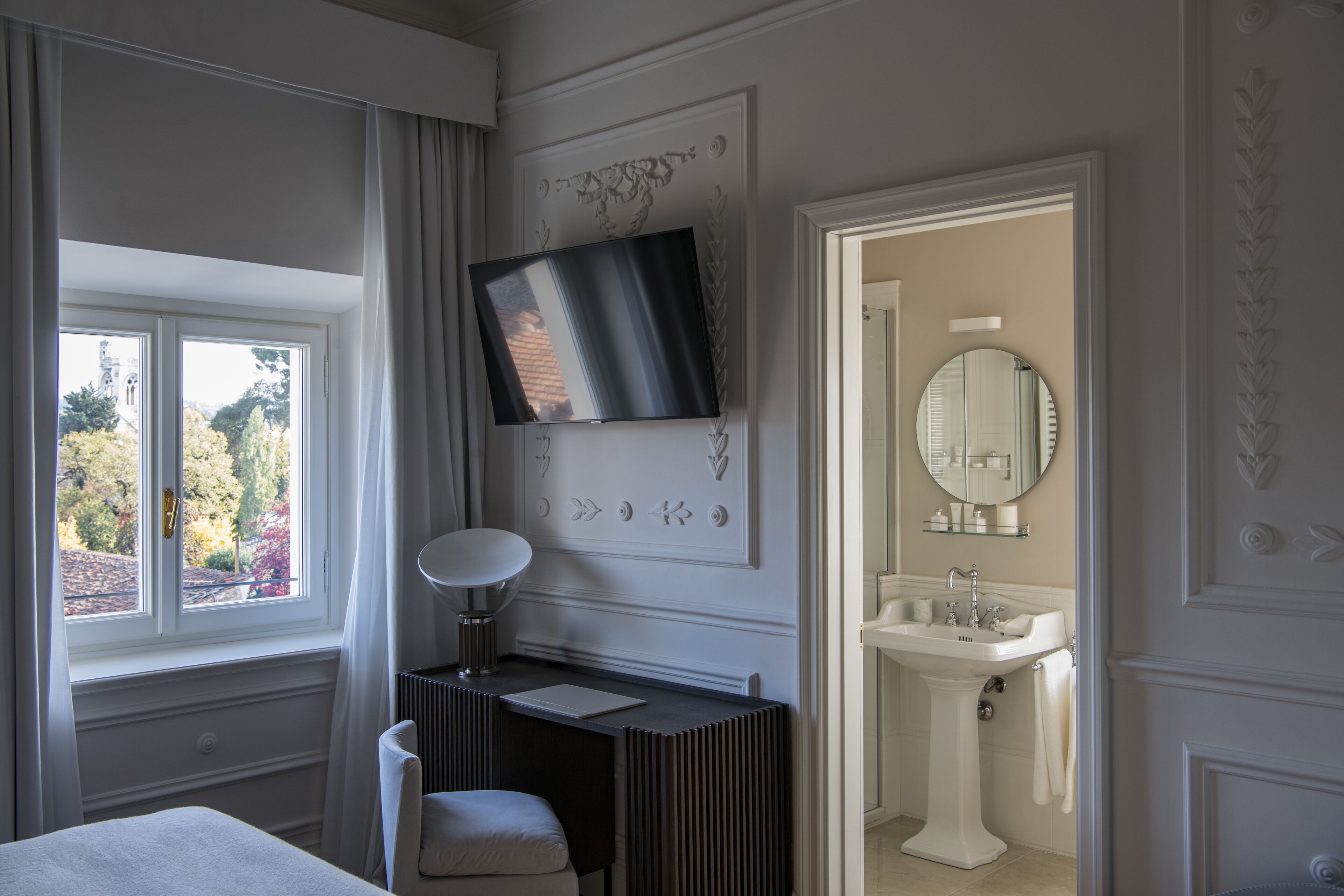 Une chambre à coucher comprenant un lit, un bureau, une télévision, une fenêtre et une salle de bain avec un lavabo et un miroir.