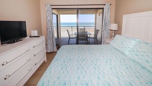 Ett hotellrum med en säng, en TV, en byrå, en balkong med utsikt över havet och ett bord med stolar.