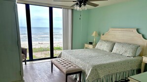 A bedroom with a large window, a ceiling fan, a bed with patterned bedding, a bedside table with a lamp, and a bench. - Sand Dollar 2-201 Direct Oceanfront Corner Unit (Saint Augustine)