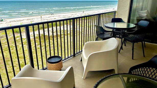 A balcony with a view of the beach, white chairs, a table, and a wicker basket. - Sand Dollar 2-201 Direct Oceanfront Corner Unit (Saint Augustine)