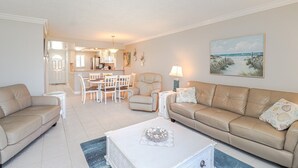 A living room with a beige sofa, a dining table with chairs, a kitchen area, and a beach-themed painting on the wall.