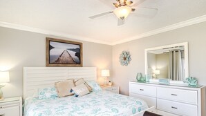 A bedroom with a bed, nightstands, a dresser, a bench, a ceiling fan, a wall-mounted mirror, and a decorative wall piece.