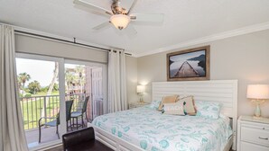 A bedroom with a bed, a bench, a nightstand, a lamp, and a view of the outdoors.