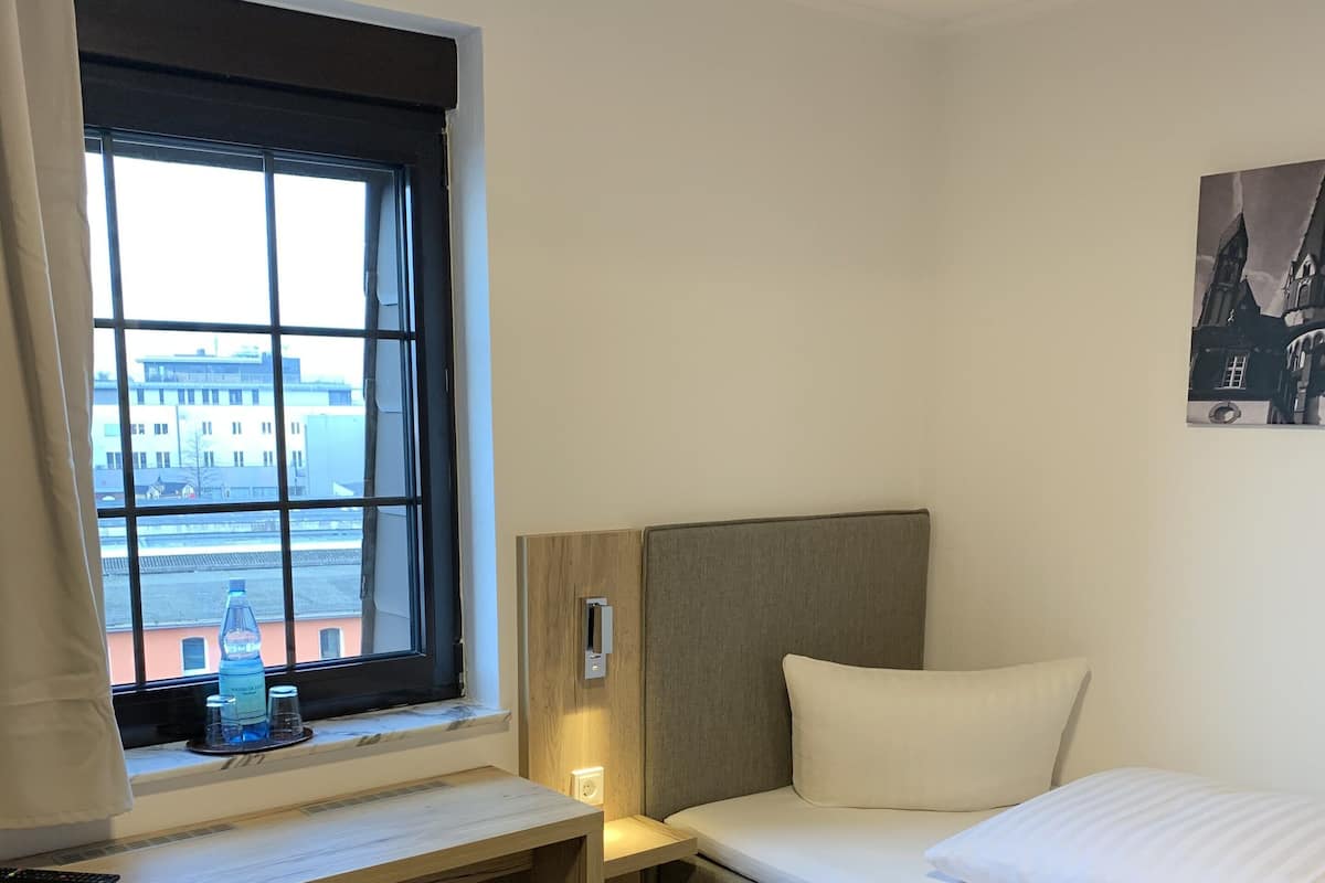 A hotel room with a bed, a desk, a chair, a window with a view of buildings, and a painting on the wall.