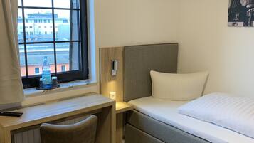 Habitación de hotel con cama, escritorio, silla, ventana con vistas a edificios y un cuadro en la pared.
