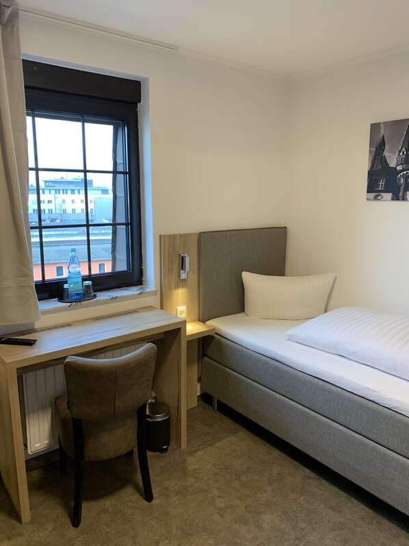 Habitación de hotel con cama, escritorio, silla, ventana con vistas a edificios y un cuadro en la pared.