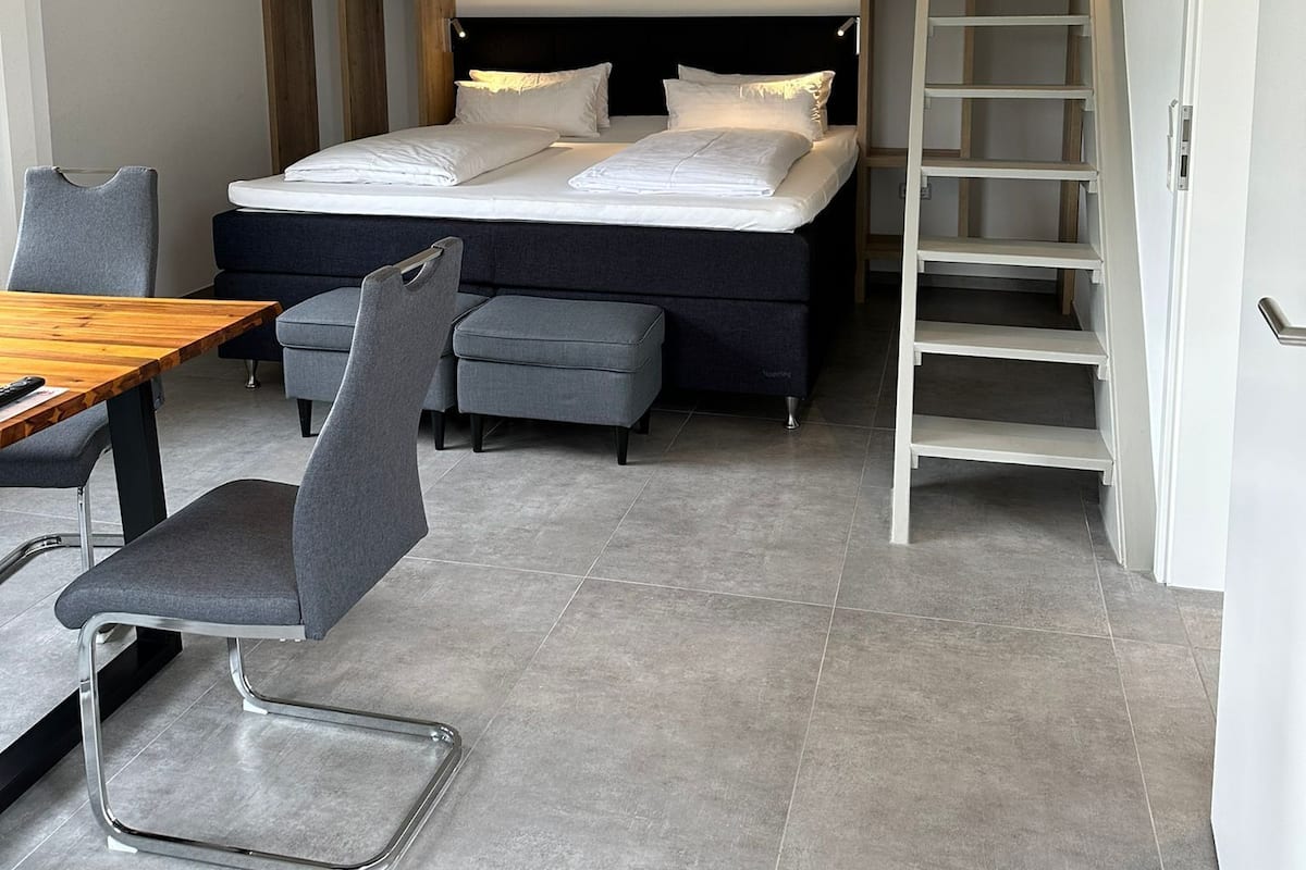 A bedroom with a loft-style bed, a dining area with a wooden table and chairs, and a staircase leading to an upper level.