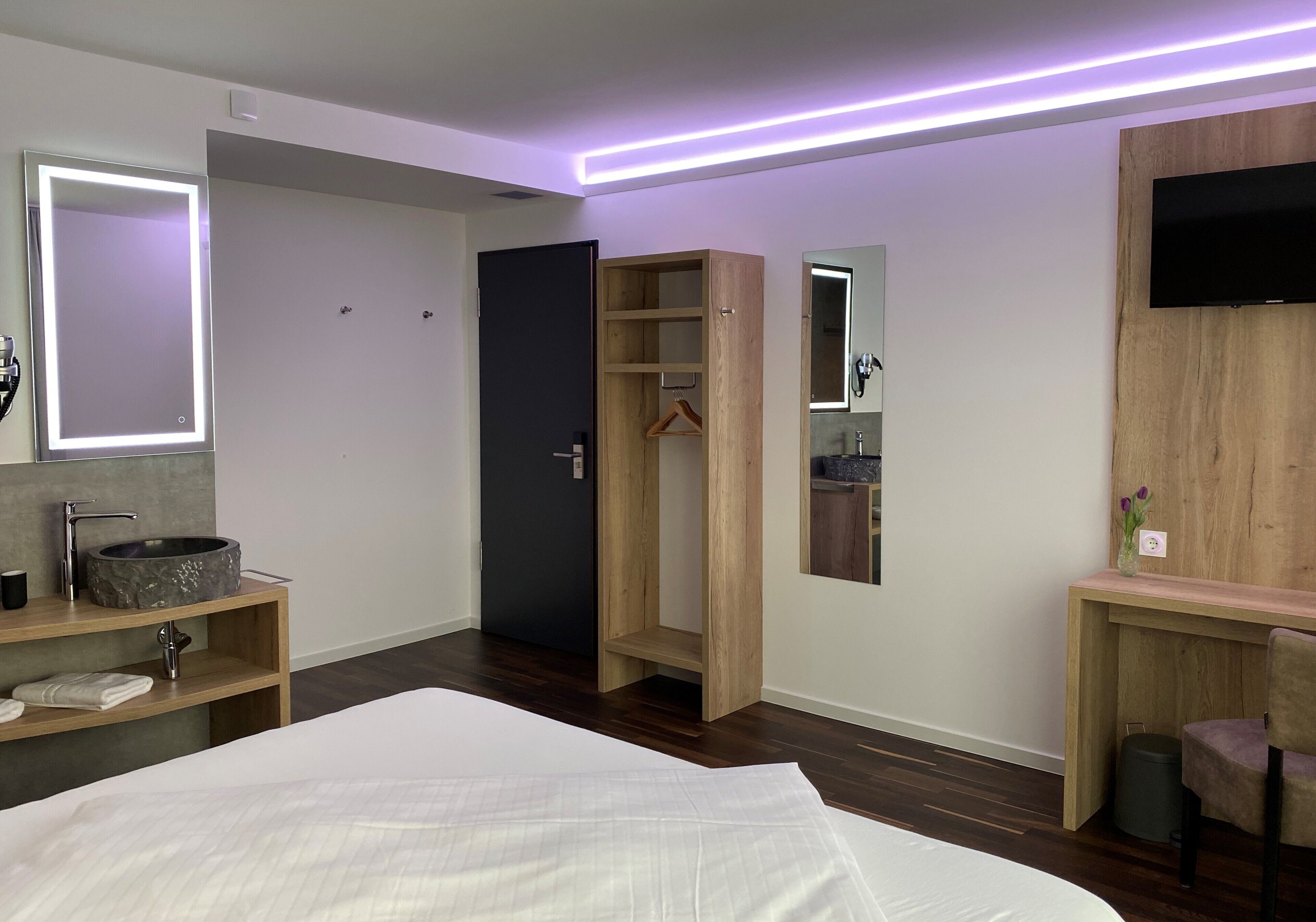 A modern bedroom with a large bed, a wooden wardrobe, a vanity with a mirror, and a TV mounted on the wall.
