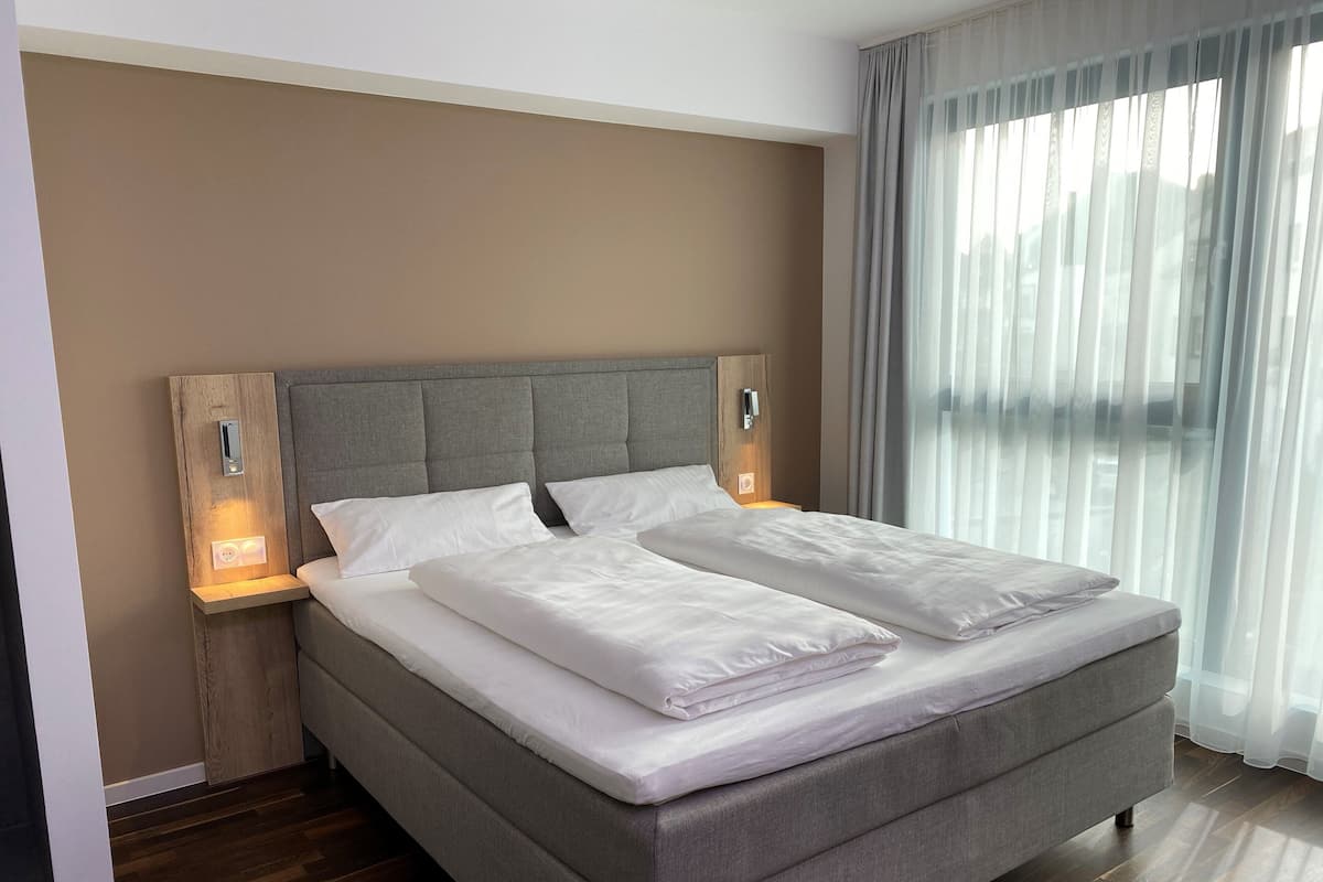 A modern hotel room with a large bed, wooden headboard, bedside lamps, and a view through sheer curtains.