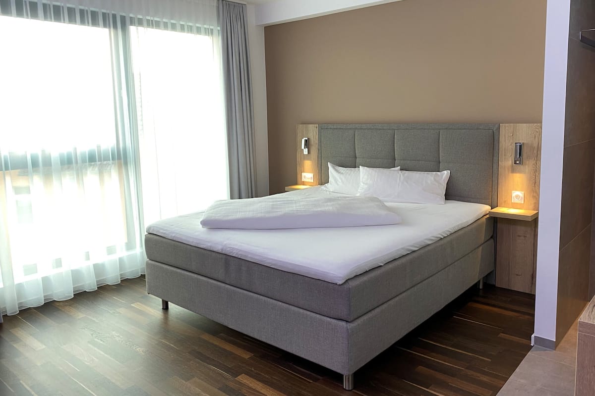 A modern hotel room with a large bed, wooden flooring, and a view of the city through large windows.